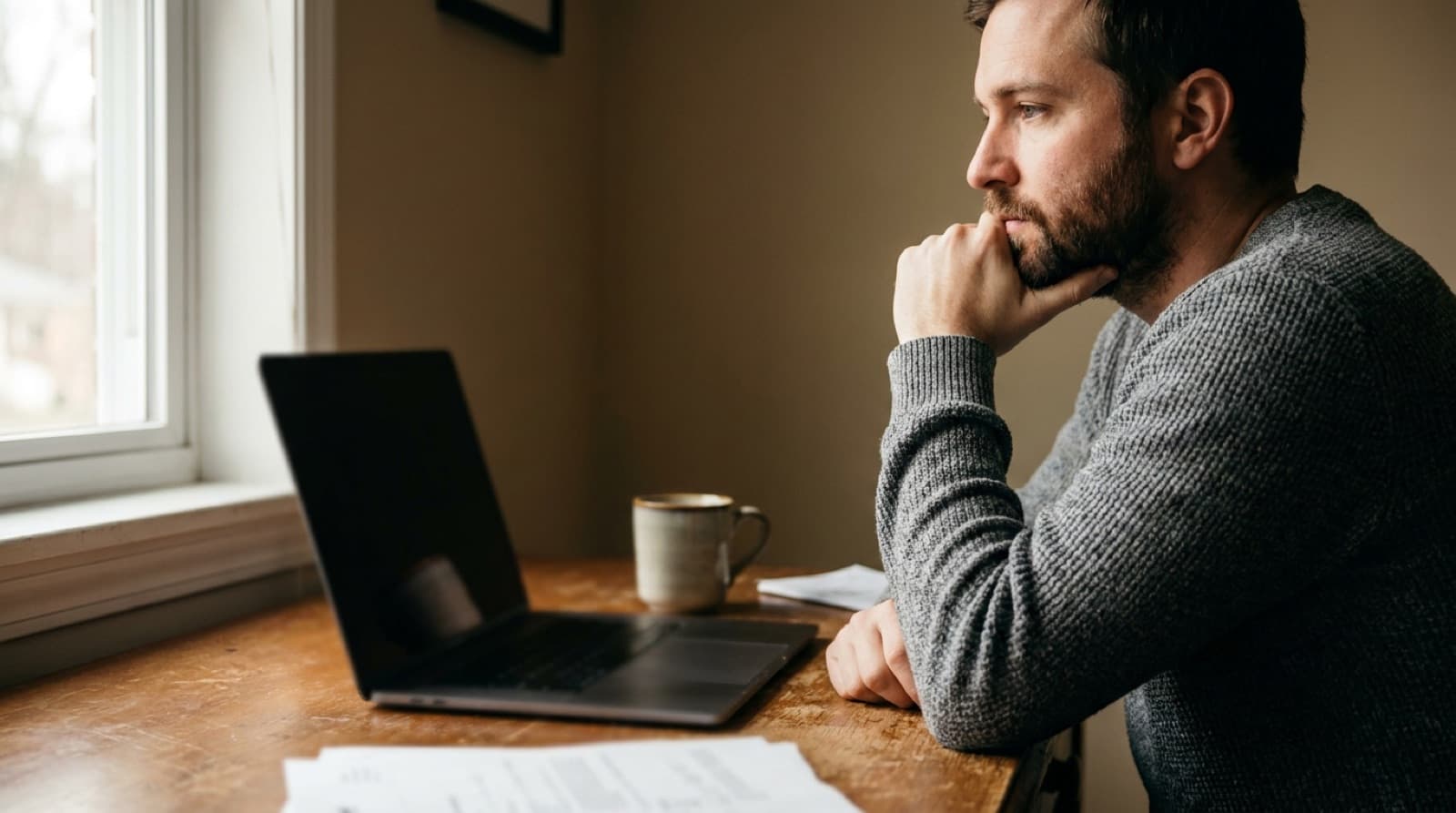 A learner at a wooden desk, mid-thought over an AI draft on a laptop, in soft morning window light